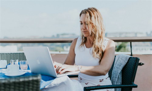 Woman working on a laptop on a sunny outdoor balcony with city view