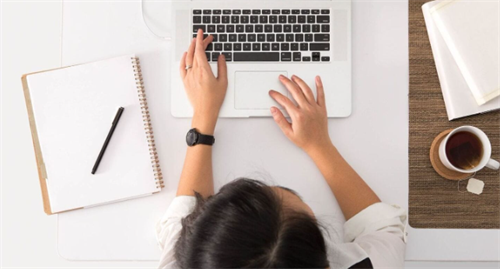 Top-down view of hands typing on a laptop at a desk with notebook and tea